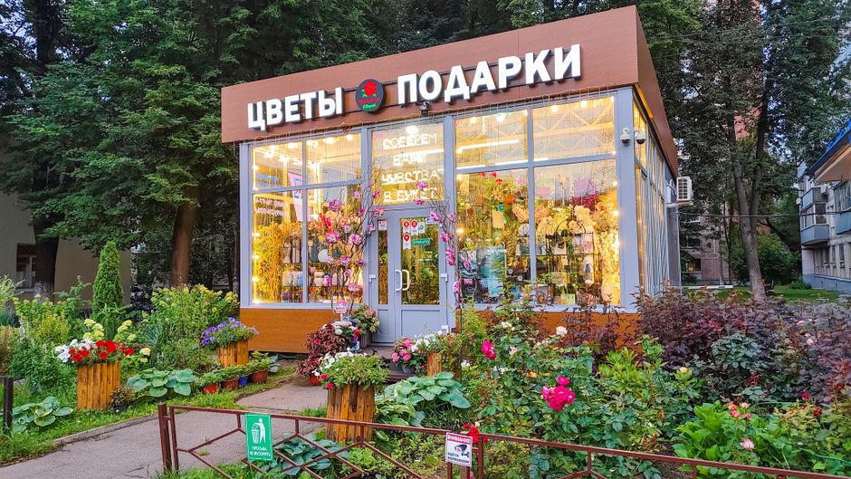 The exterior of Brent Florist, a flower shop with large glass windows displaying illuminated floral arrangements and gifts inside. The shop has a modern facade with a brown frame and signage with Cyri