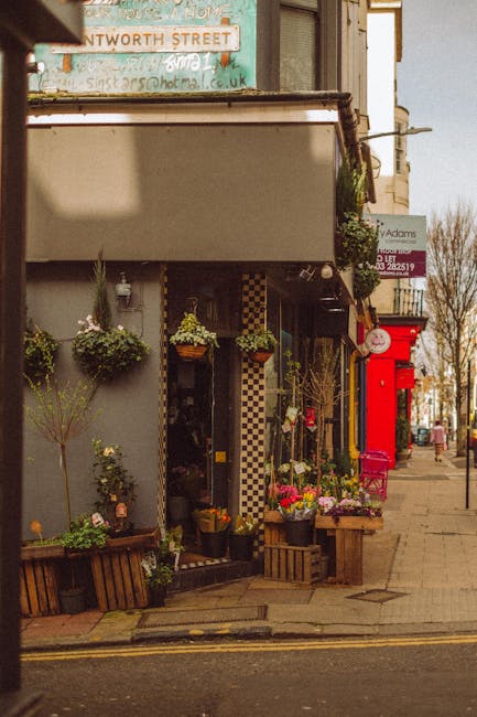 The image depicts the exterior of a charming flower shop with a welcoming display of fresh floral arrangements outside. The arrangement includes vibrant pink, yellow, and white flowers in pots and bas