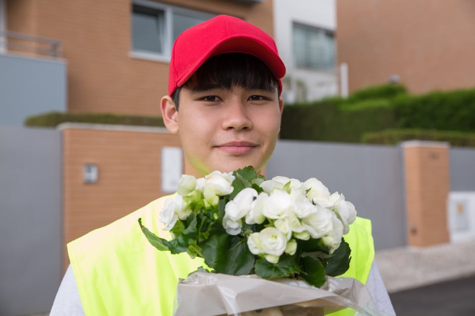 A young delivery person wearing a red cap and neon yellow high-visibility vest holds a bouquet of fresh white roses wrapped in white paper, standing outdoors in an urban residential area with modern h