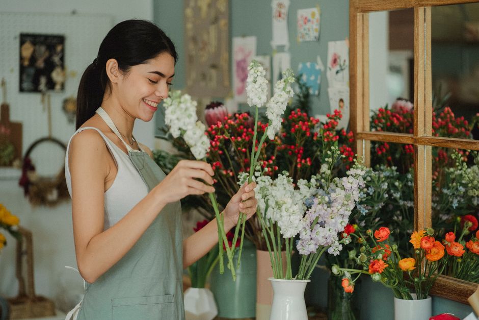 A young woman with dark hair tied back, smiling gently as she arranges a bouquet of white lilacs and other mixed flowers with green stems. She is wearing a light-colored apron over a sleeveless top an