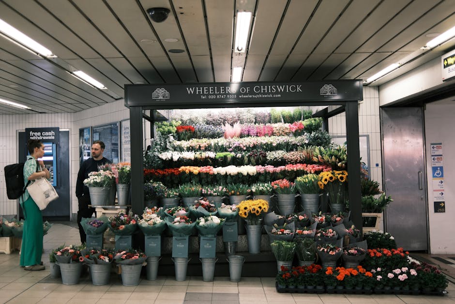 A vibrant flower stall at Kilburn station featuring an array of fresh floral bouquets arranged on multiple levels. The display includes sunflowers with bright yellow petals, various roses in pink, red