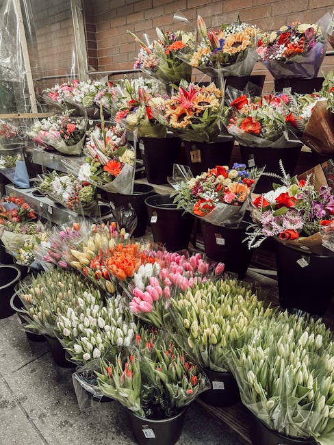 An outdoor display of an extensive assortment of fresh flowers arranged in black buckets and wrapped in decorative paper, including pink and white tulips, orange lilies, and mixed bouquets of roses an