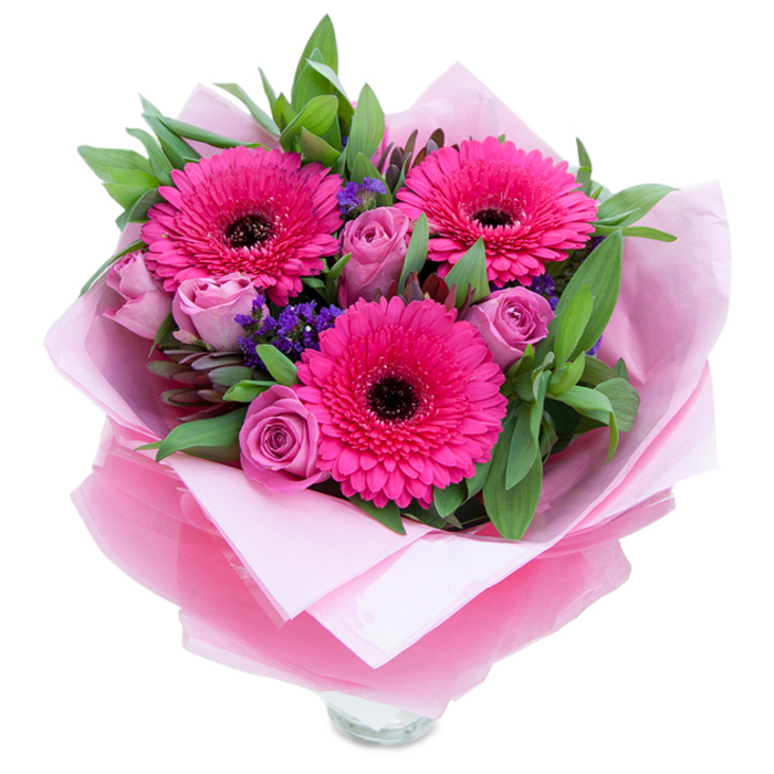 I picture this bouquet as a tidy, joyful surprise: three striking fuchsia Gerbera daisies dominate the scene, their broad, radiating petals and pronounced dark centres creating a vivid, cheerful focal point. Around them, roses in a soft lavender-pink or dusty rose tone open in layered, velvety whorls, offering moments of tenderness and quiet romance amid the louder gerbera statement. Small sprays of purple statice or limonium are woven through the stems, their papery clusters adding a textural spark and a cool blue-violet note that balances the warm pinks. The greenery-full, glossy leaves that read as salal or eucalyptus-wraps the bouquet in a deep, verdant hush, giving the composition structure and a freshly gathered-from-the-garden feel. The florist's wrapping is carefully visible: an inner cushion of matte pale-pink tissue paper that creates a soft base, topped with a slightly crinkled, translucent fuchsia-pink cellophane that shimmers and ties the colour story together. The lower edge of the image reveals the top rim of a clear glass vase, suggesting the bouquet is intended for immediate display and the tactile comfort of clean water and glass. The aroma is understated: warm rose perfume with a whisper of green sweetness and the faint, dry note of statice. This arrangement feels versatile - equally suited to a celebratory birthday table in a Willesden home, a thank-you left on a neighbour's doorstep, or a bright, reassuring gift to mark an anniversary - a polished, heartfelt offering from a local florist who understands everyday moments of joy.
